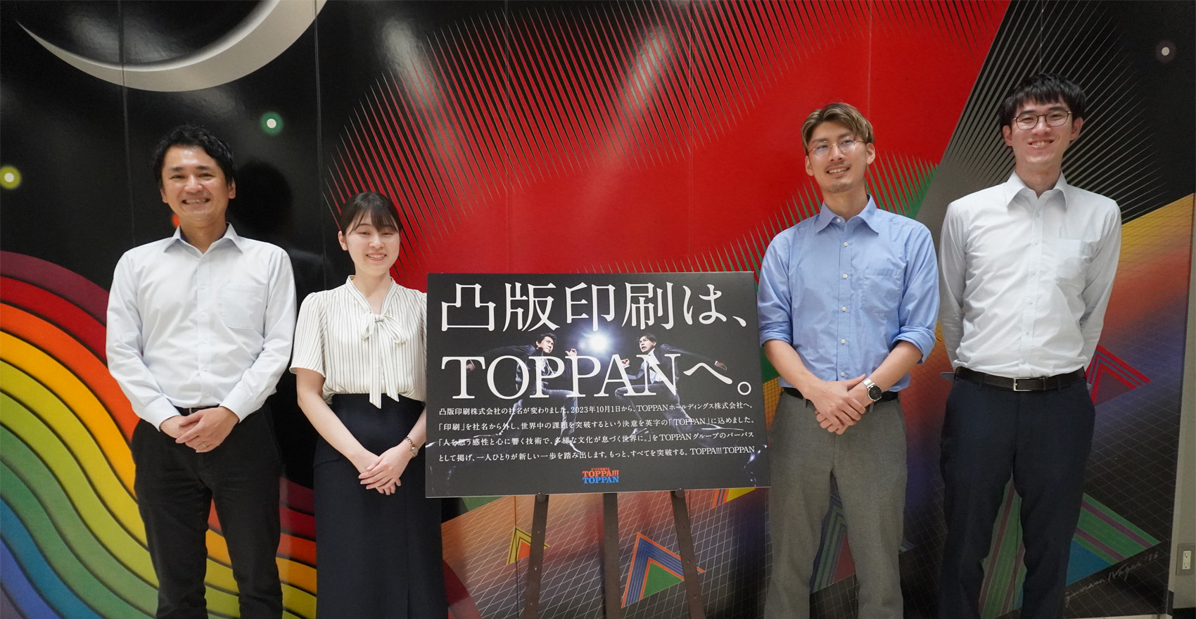 男性社員3名と女性社員1名が「凸版印刷は、TOPPANへ」の看板とともに横一列に並び、笑顔をむける様子