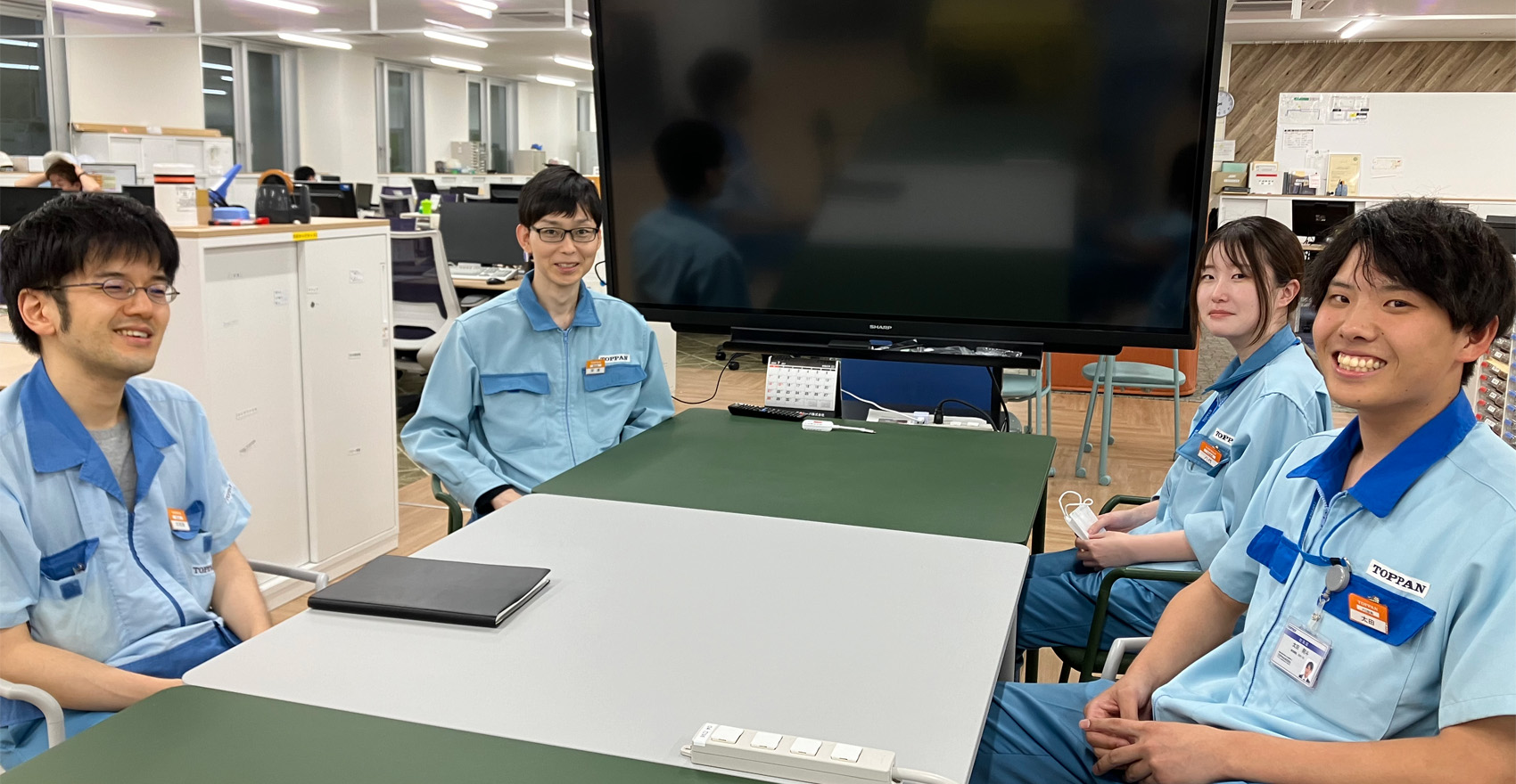 男性社3名と女性社員1名がデスクで互いに向かい合って座っており、こちらに笑顔をむける様子